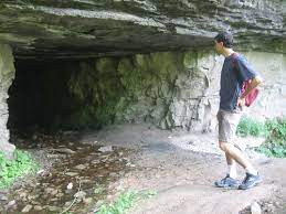 man checking out the tunnel, rocky and dry floor, blue shity, red bag, crocs, wild plants